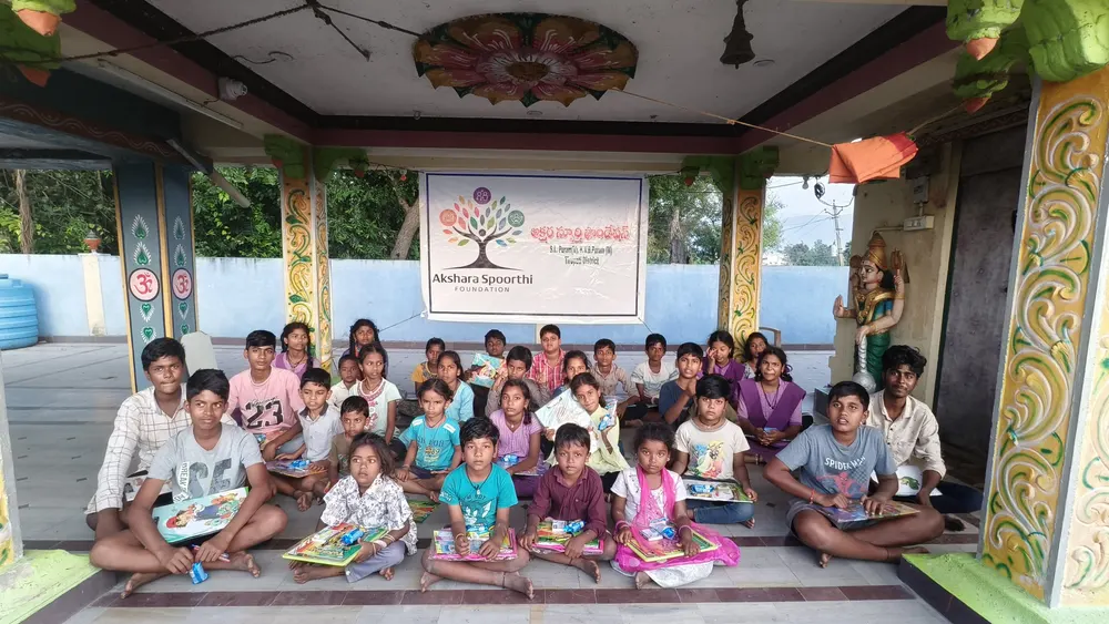 About thirty children sit cross-legged with new books under a painted pavilion, an AKSF banner behind them