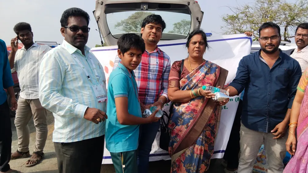 Woman in a saree hands water and a food packet to a boy as men stand by a car with the foundation banner